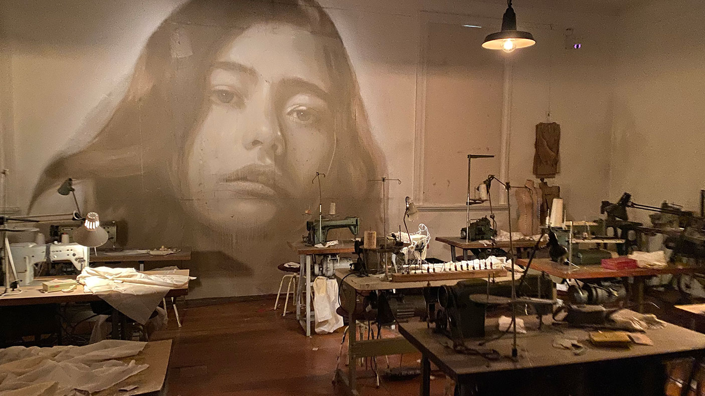 A scene from the installation TIME • RONE showing a commerical sewing room, with a young woman's portrait on the wall.