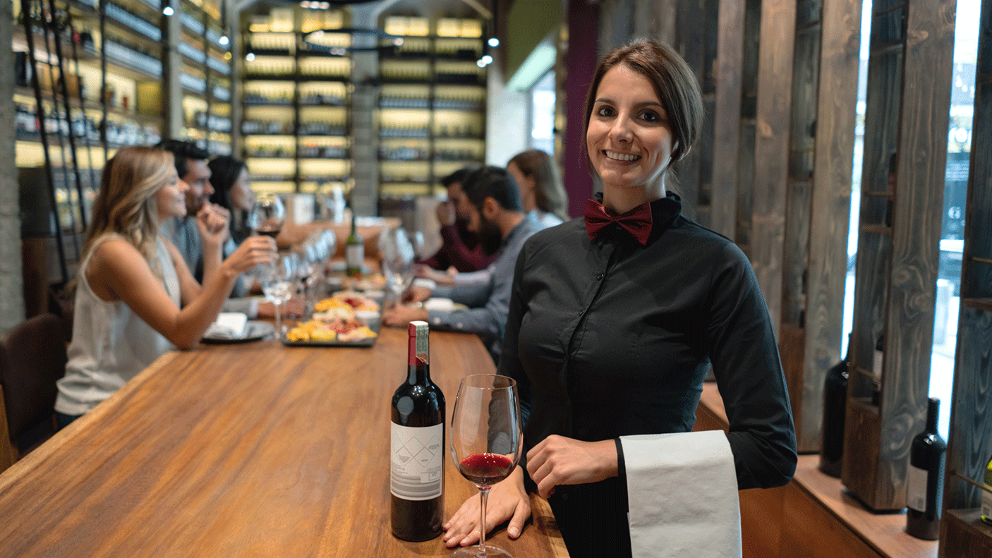 A waiter with a bottle of red wine and a glass, stands near a table of people in a bar, who are eating food and drinking.