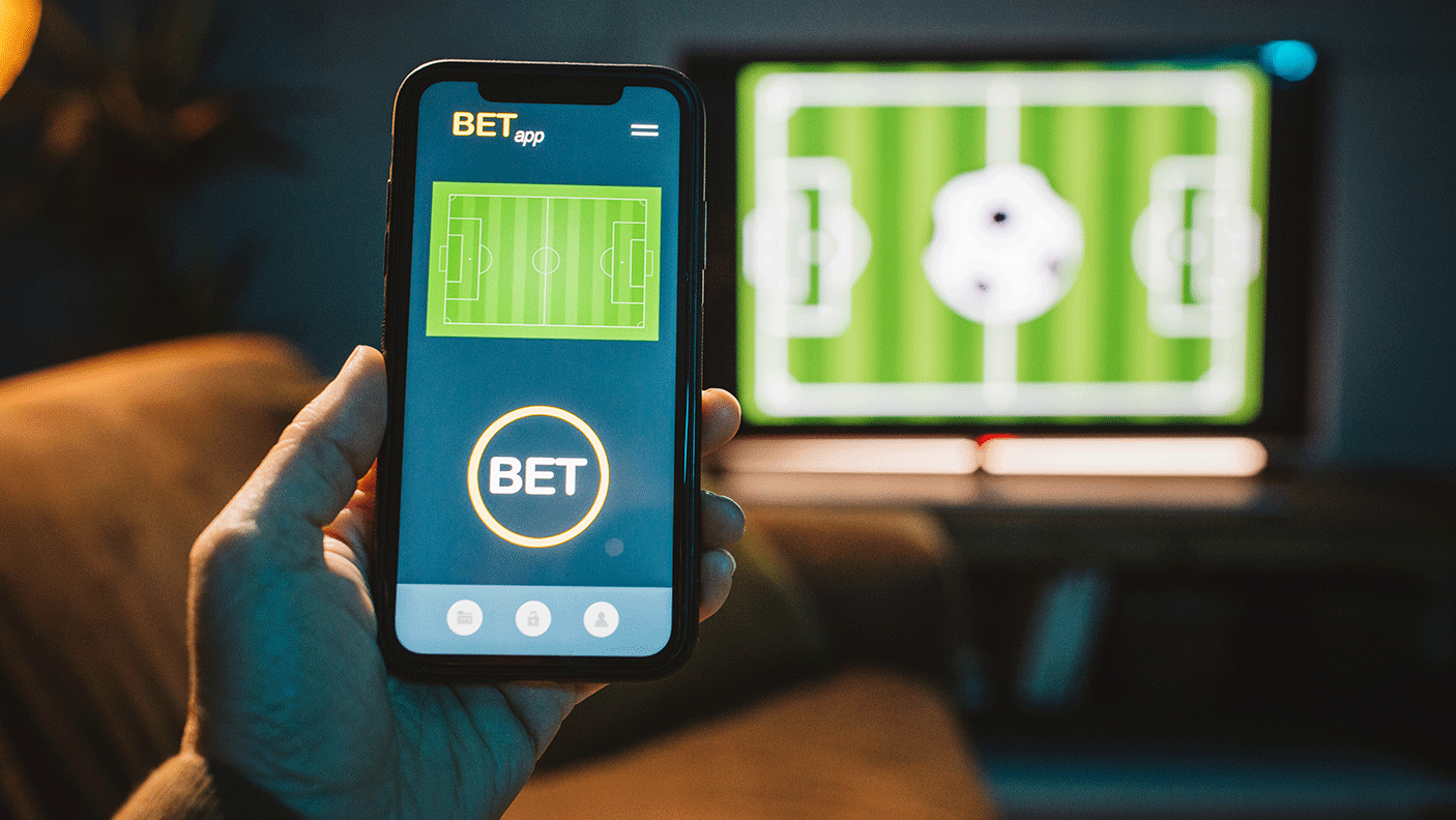 A person holds a mobile phone with a sports betting app open on the screen.  In the backgrounds is a TV with a sporting game on
