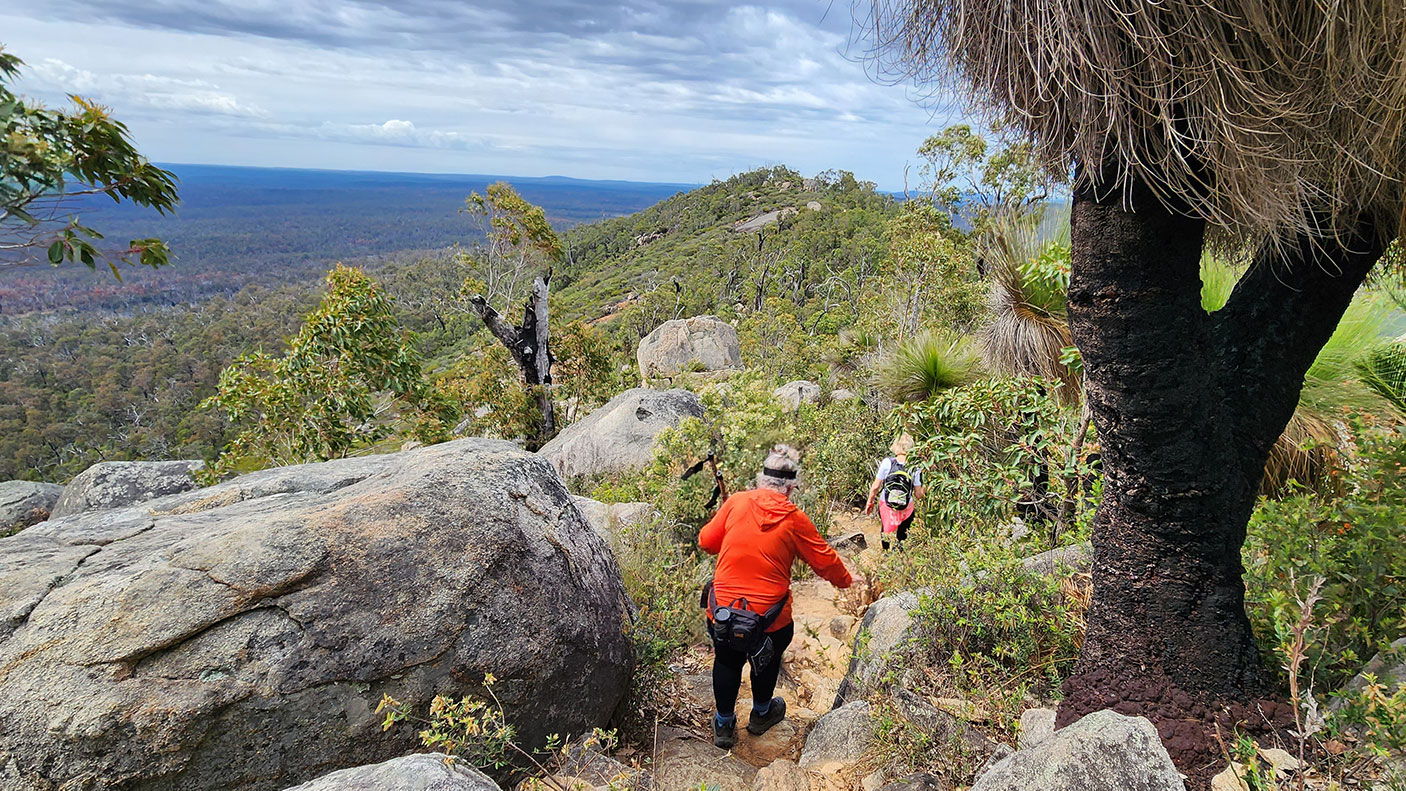 Two people hiking in the Perth hills, next to a grass tree and granite boulders